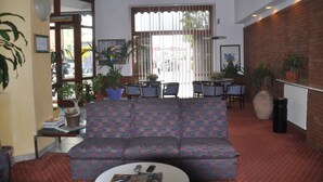 Una sala de estar con un sofá estampado, una mesa de centro y una zona de comedor con sillas y una mesa. Hay plantas en macetas y un radiador en la pared.
