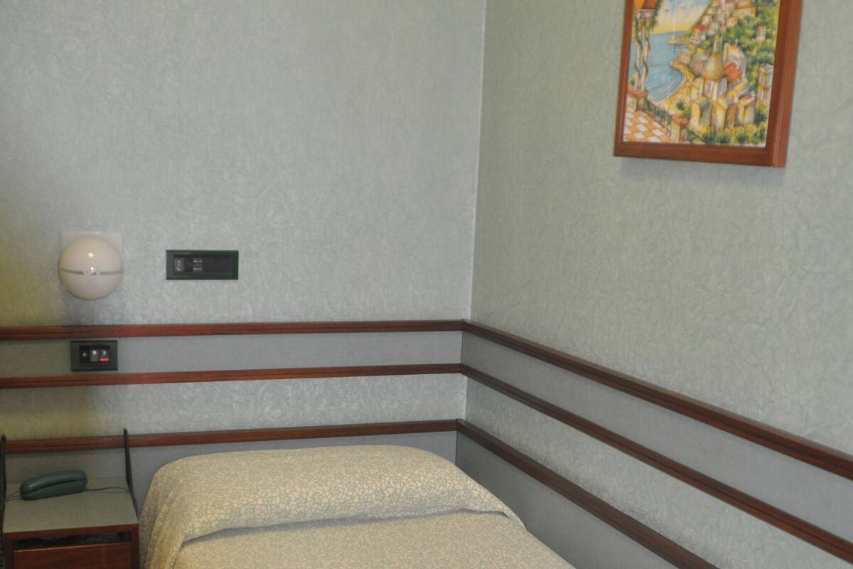 A single bed with a patterned bedspread, a bedside table with a lamp, and a painting on the wall.