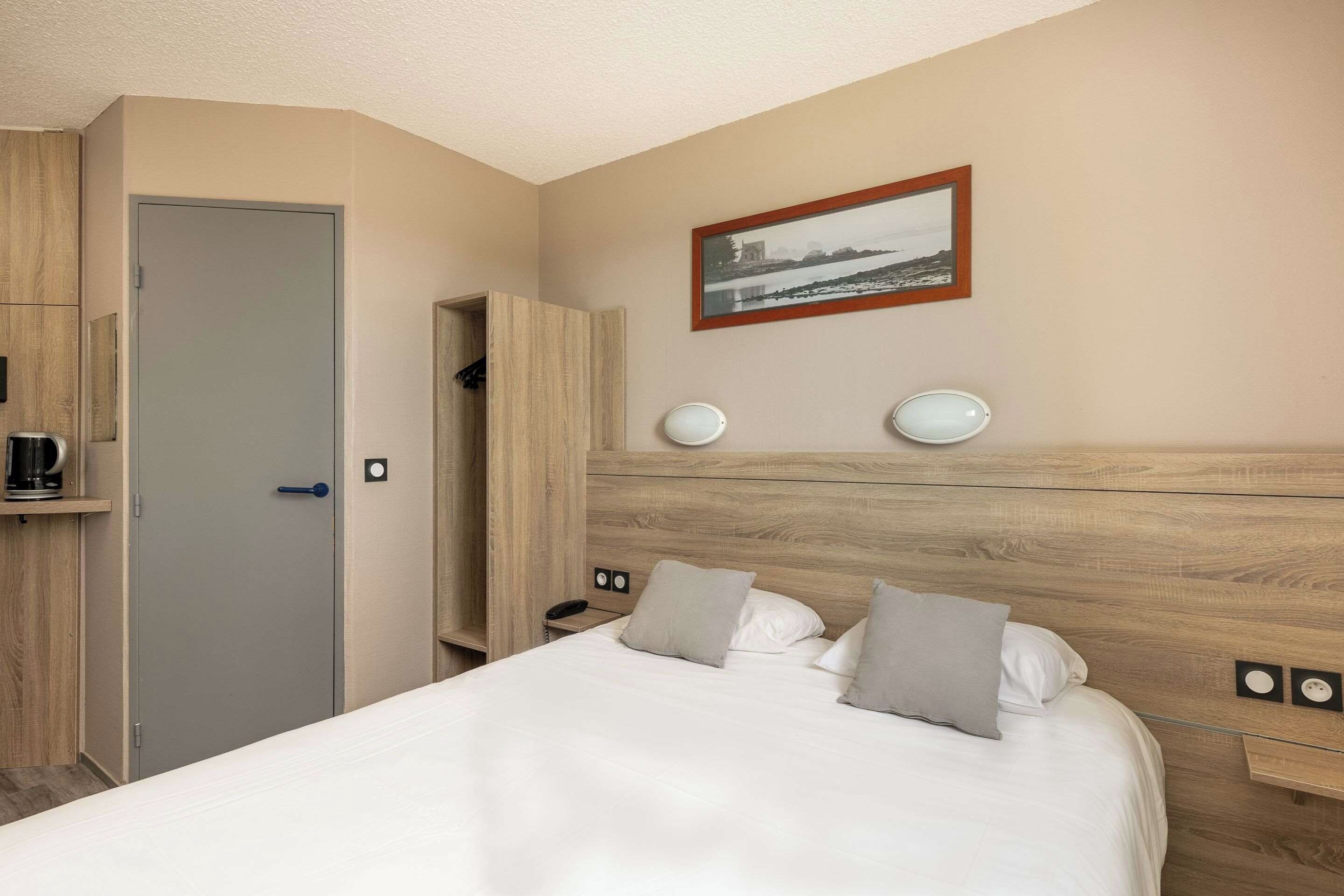 A hotel room with a bed, a bedside table, a nightstand, a coffee machine, a wardrobe, and a framed picture on the wall.