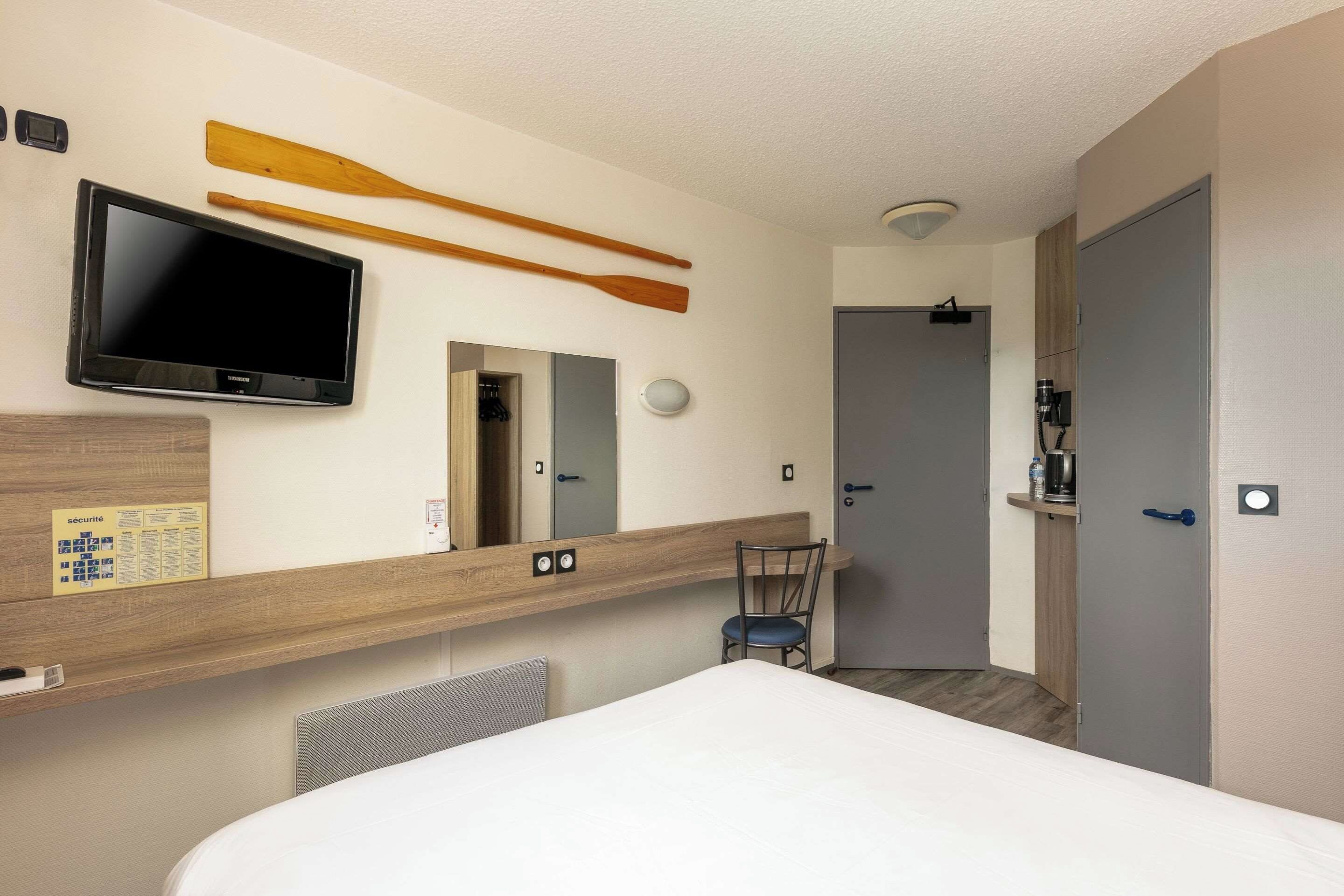 A hotel room with a bed, a TV, a mirror, a wooden shelf, a chair, a wardrobe, and a door.