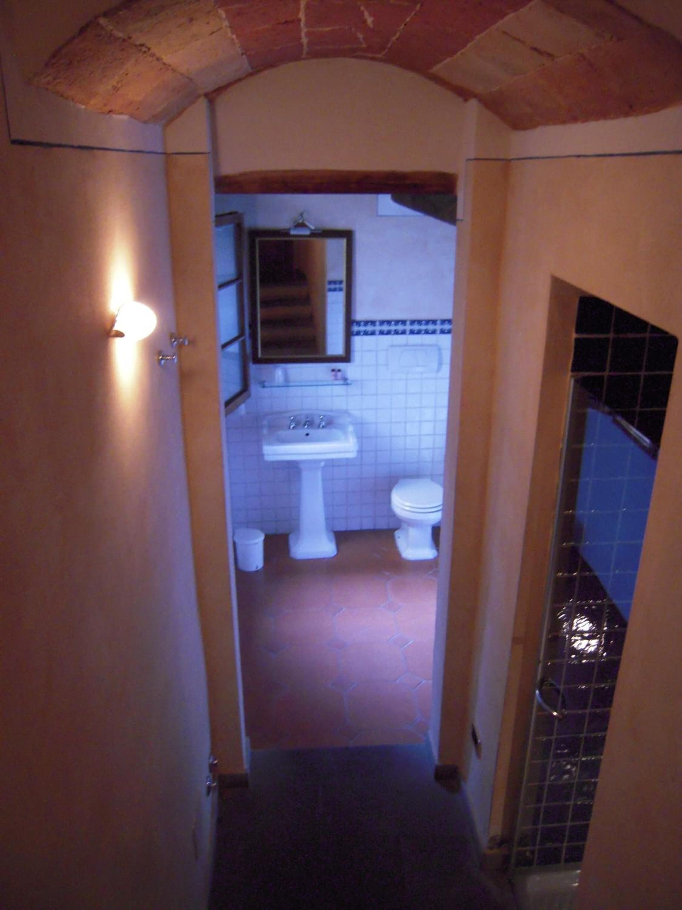 A narrow hallway leading to a bathroom with a sink, two toilets, and a mirror.