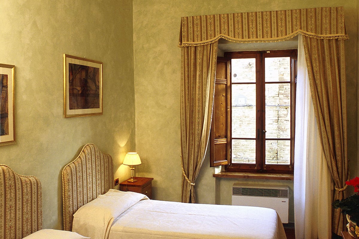 A hotel room with two beds, a window with curtains, a wall-mounted picture, and a bedside table with a lamp.