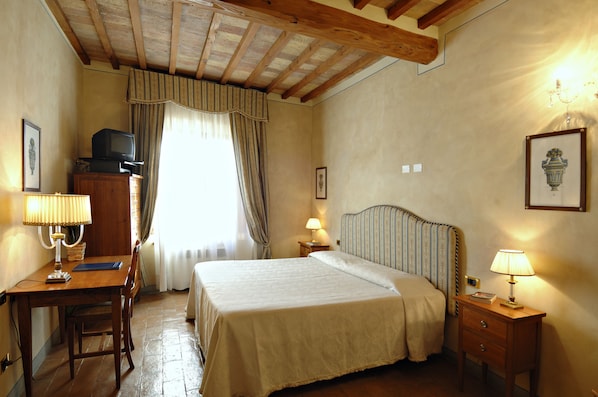 A hotel room with a wooden desk, a bed with a striped headboard, a nightstand with a lamp, and a window with curtains. - Palazzo al Torrione 2 (San Gimignano)