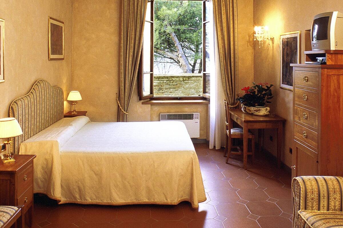 A hotel room with a bed, a desk with a television, a chair, a window with curtains, and a wall with decorative molding.