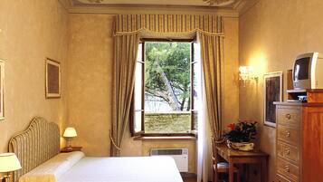 Una habitación de hotel con cama, escritorio con televisor, silla, ventana con cortinas y un muro con molduras decorativas.