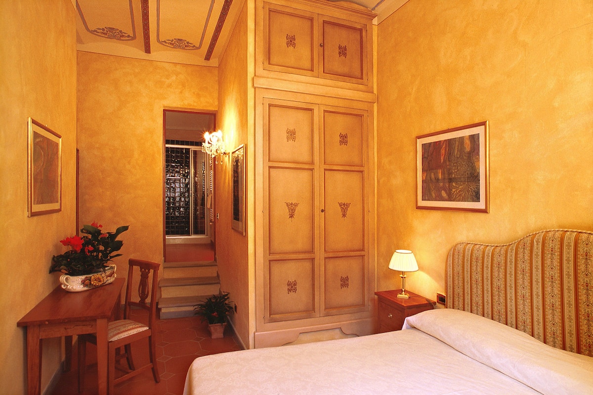 A hotel room with a bed, a desk with a chair, a potted plant, a wardrobe, and a painting on the wall.