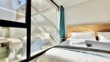 Una habitación de hotel con un ventanal grande, un cabecero de madera, una cama bien hecha con sábanas blancas y una mesita de noche con una lámpara.