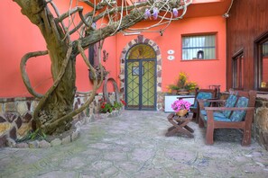 Un patio con piso de piedra, una mesa de madera con sillas, un árbol grande y un edificio con pared roja y puerta verde.