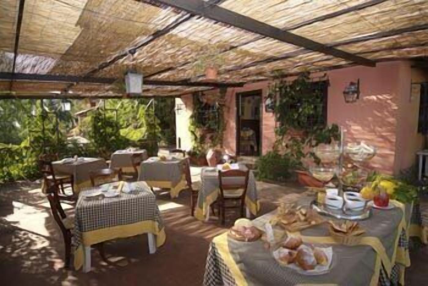 An outdoor patio with tables and chairs, covered by a thatched roof, surrounded by greenery and a pink building.