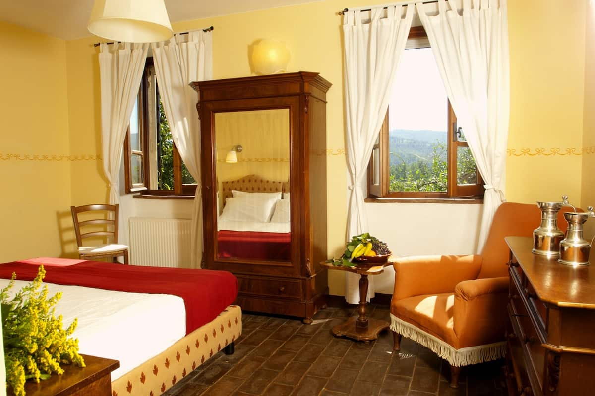 Une pièce comprenant un lit, une armoire en bois, une chaise, une table avec un panier de fruits et une vue sur la verdure par la fenêtre.