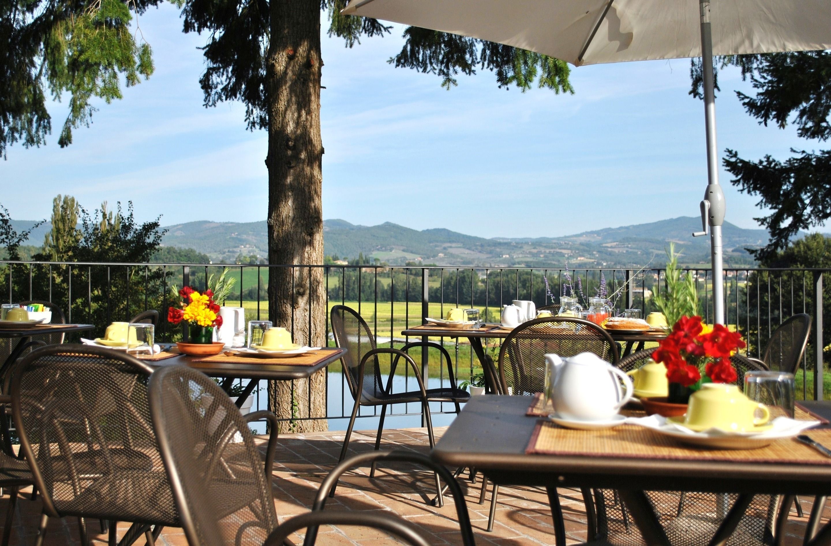 Un espacio de comedor al aire libre con mesas y sillas, vistas a un lago y colinas, y un árbol que da sombra.