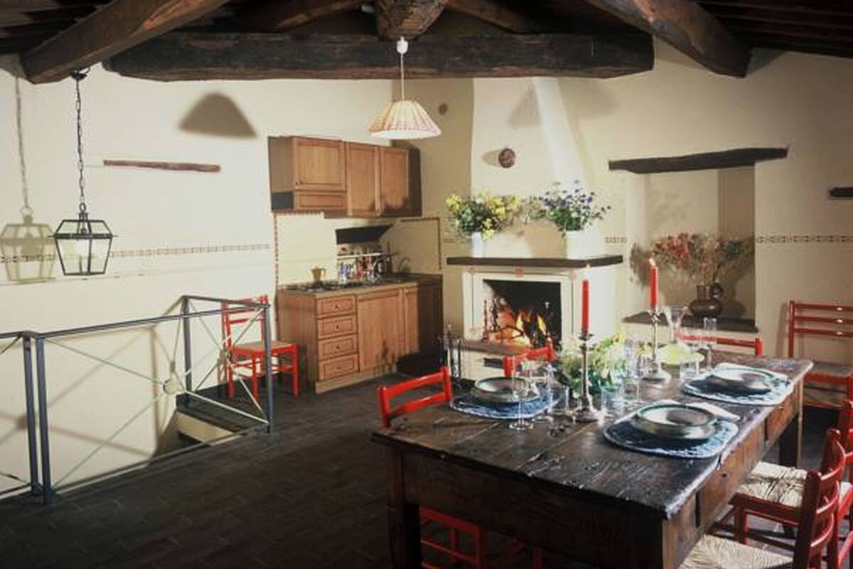 Un coin repas avec une table en bois dressée pour le repas, des chaises en bois, un luminaire suspendu et une cuisine en arrière-plan.
