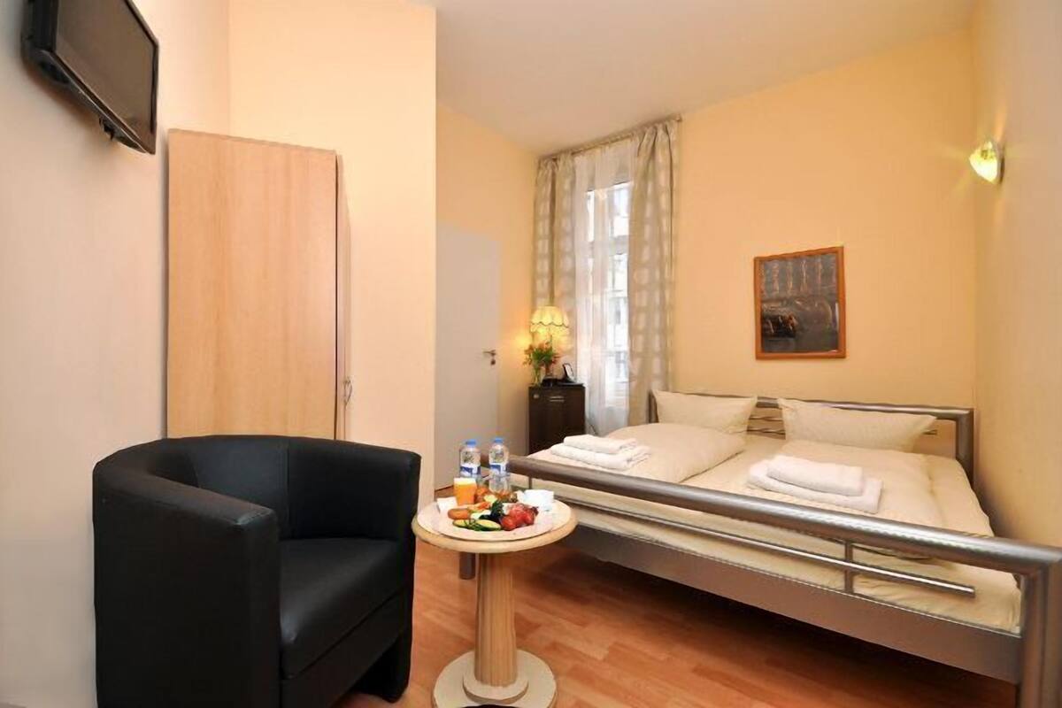 Une chambre d’hôtel avec un lit, une petite table avec le petit-déjeuner, un fauteuil noir et un téléviseur fixé au mur.