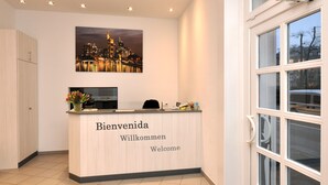 A reception desk with a cityscape painting above it, a vase with flowers, and a view of the street through the window. - Apadana Hotel (FRANKFURT AM MAIN)