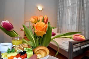 A breakfast setup with croissants, fruits, and flowers on a table in a room with a bed and a window.