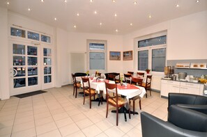 A dining area with multiple tables and chairs, a kitchen area with cabinets and appliances, and a view of the outside through large windows. - Apadana Hotel (FRANKFURT AM MAIN)