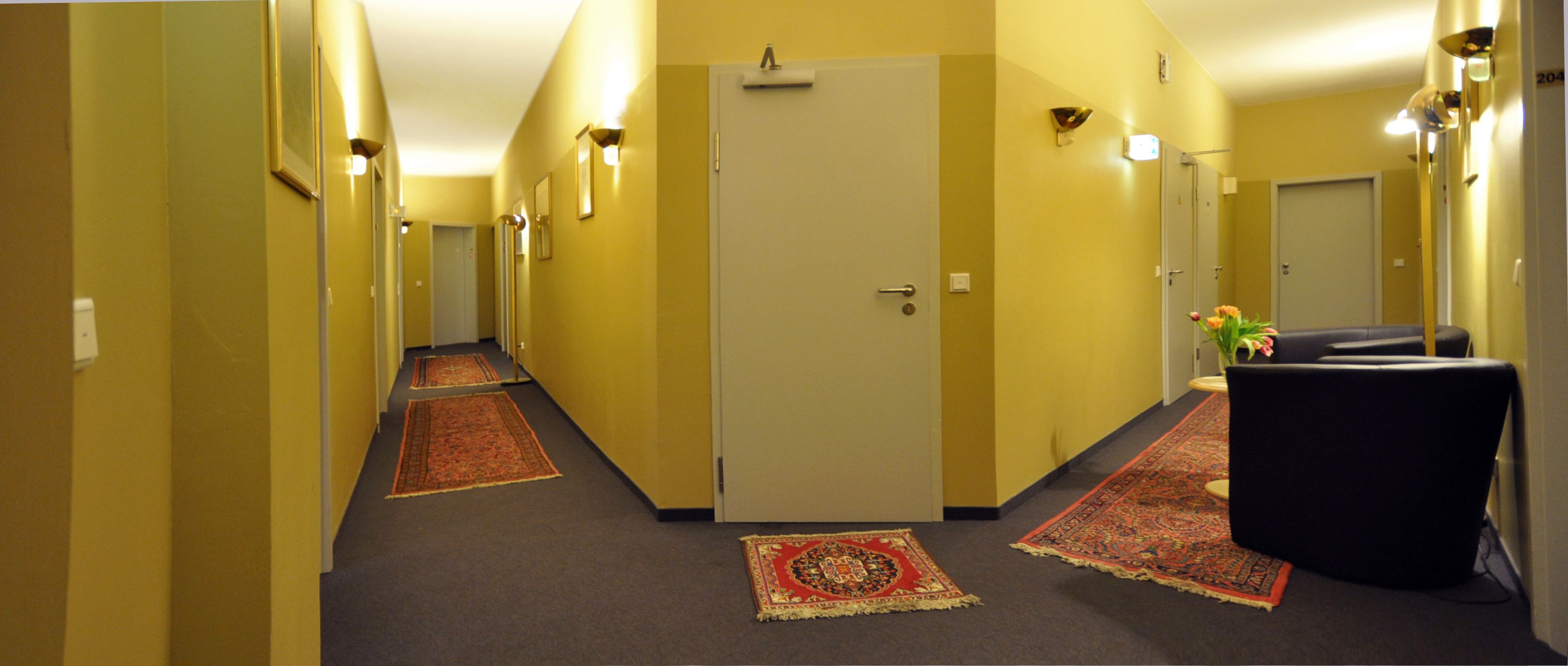 A hotel hallway with carpeted floors, a door at the end, and a black chair with a vase of flowers.