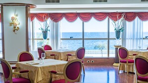 A dining area with tables set for four, large windows with ocean views, and red curtains. - Hotel Palace (Catanzaro)