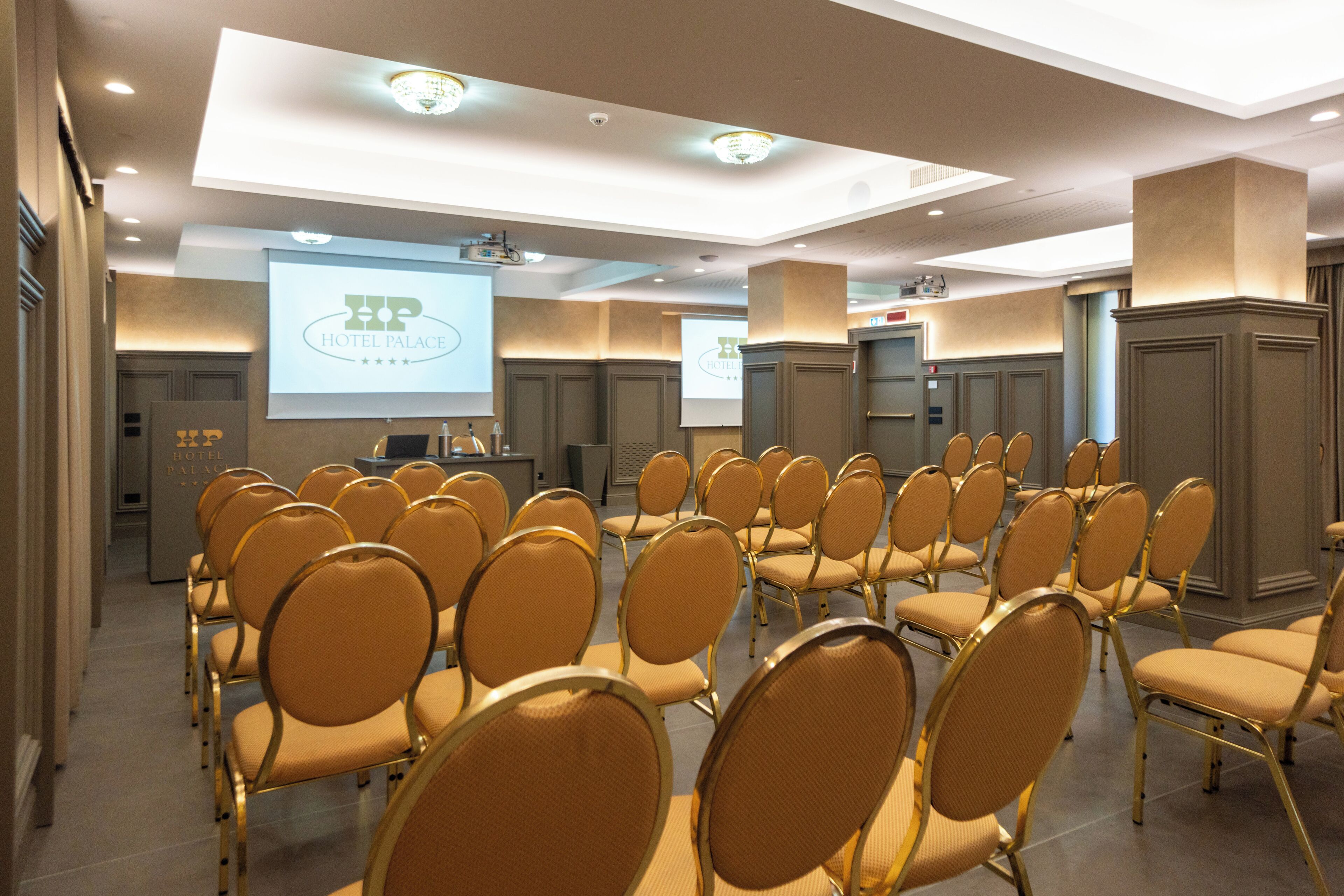 Un salón de conferencias con hileras de sillas, un atril y una pantalla que muestra 'HOTEL PALACE'.