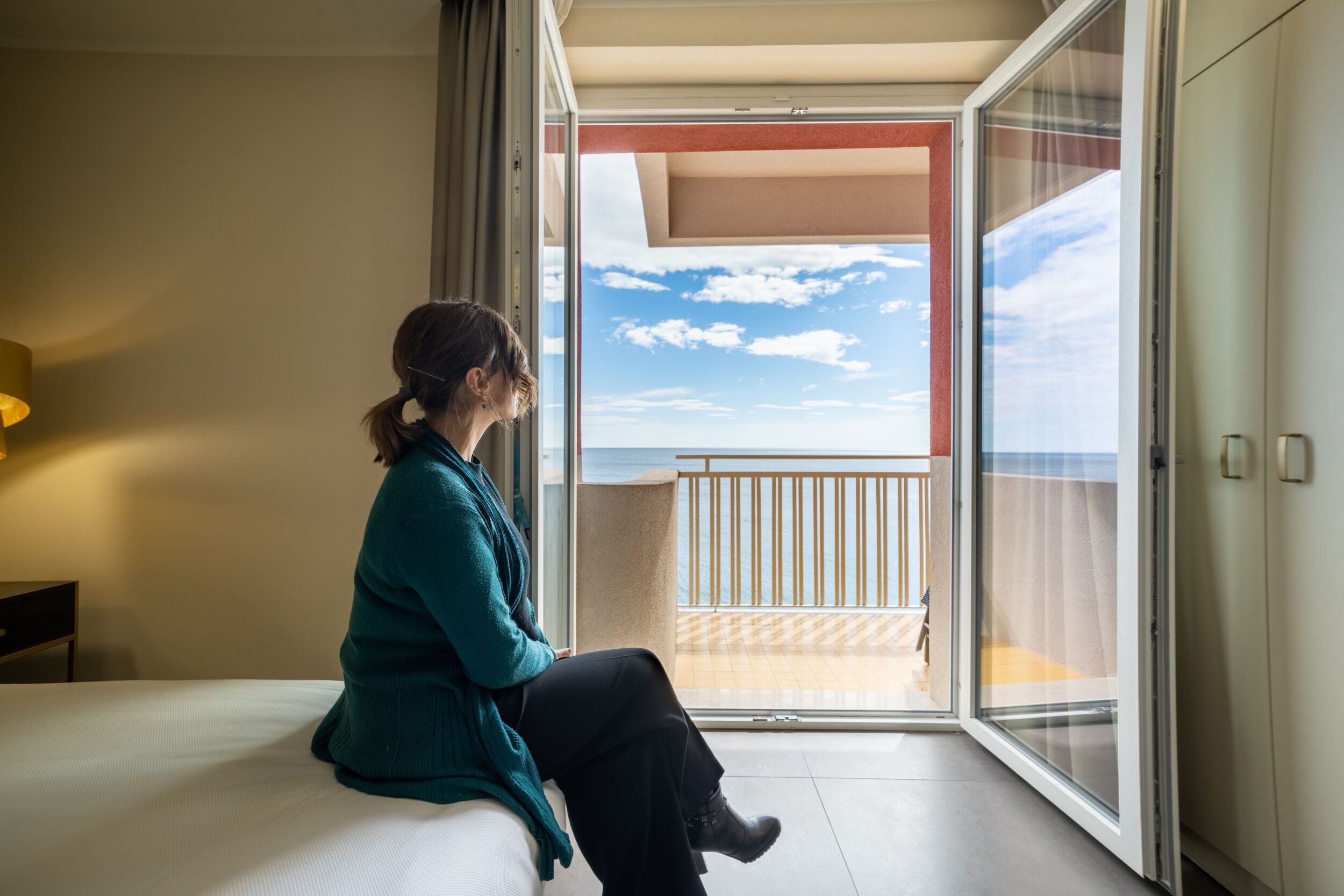 Una persona seduta su un letto in una stanza con un'ampia porta-finestra scorrevole che si apre su un balcone con vista sul cielo e sulle nuvole.