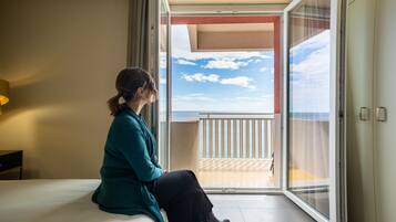 Una persona seduta su un letto in una stanza con un'ampia porta-finestra scorrevole che si apre su un balcone con vista sul cielo e sulle nuvole.