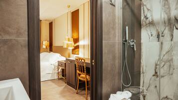 Un baño de hotel moderno con ducha, inodoro y lavabo. Hay una silla de madera y un escritorio con lámpara.