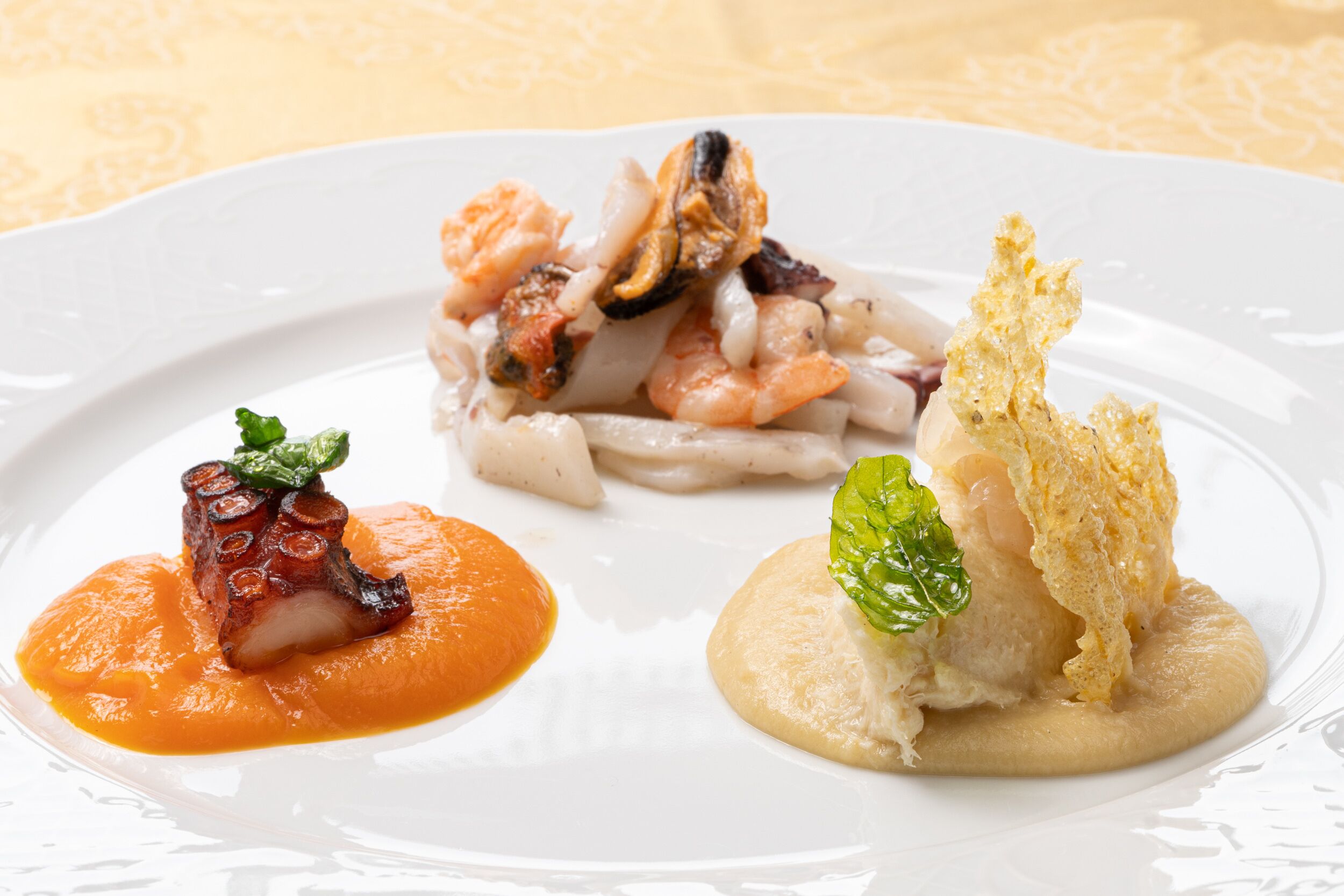 Une assiette blanche contenant trois plats de fruits de mer, chacun accompagné d’une sauce et d’une garniture différentes.