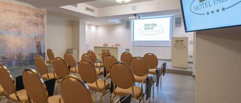 A conference room with rows of chairs, a projector screen displaying 'Hotel Palace', and a large abstract painting on the wall.