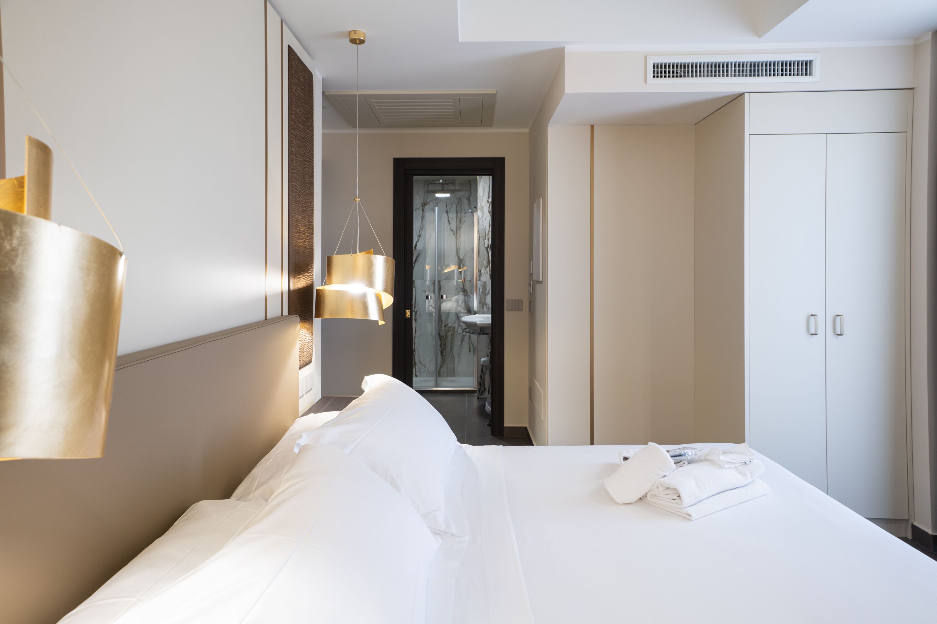 Habitación de hotel con una cama grande, una lámpara colgante, un armario y un baño visible a través de una puerta de vidrio.