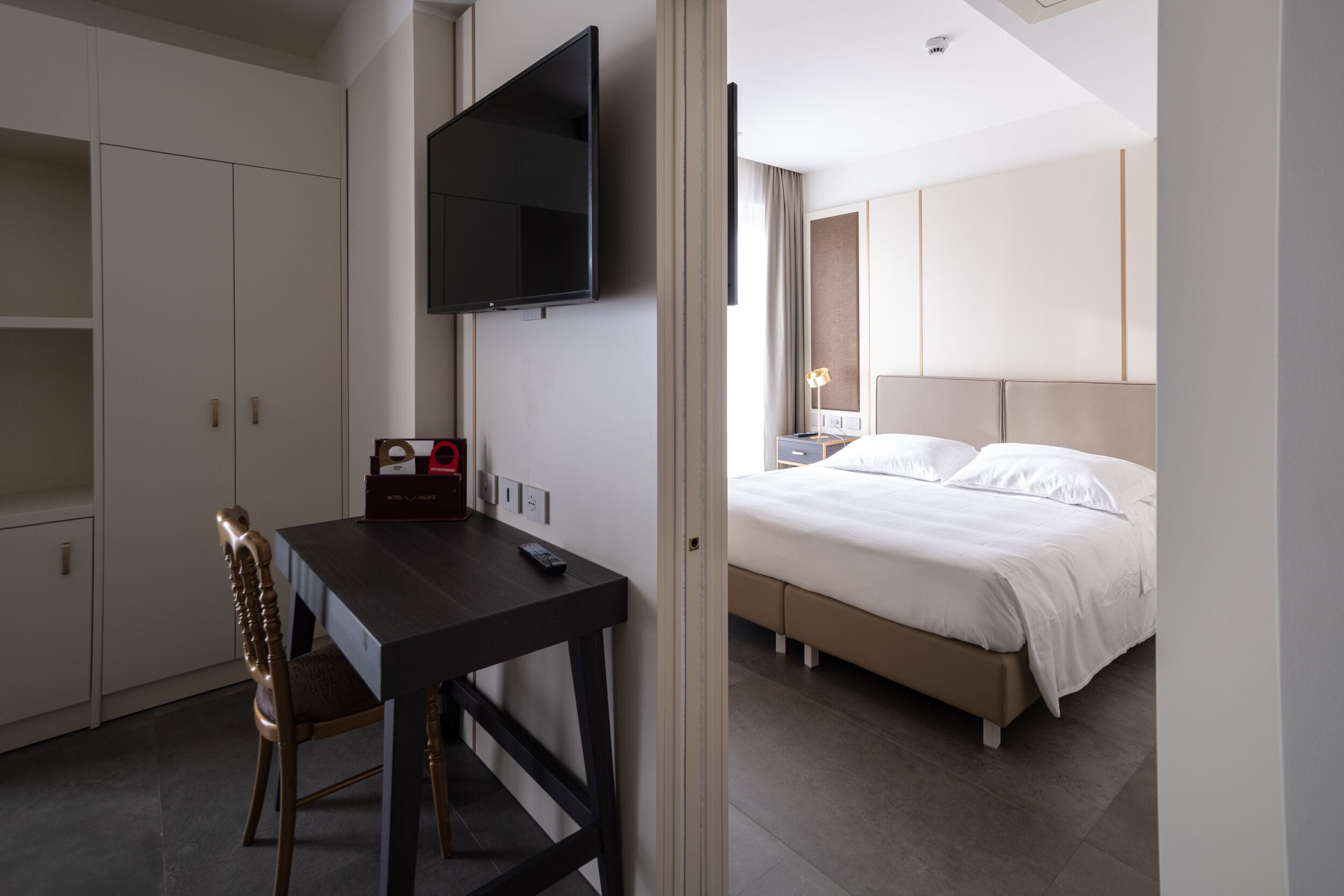 Una habitación de hotel con cama, escritorio, silla, televisor montado en la pared y armario.