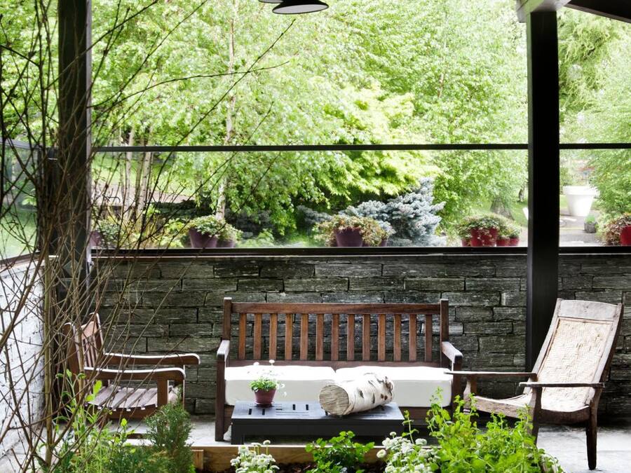 A covered outdoor seating area with a bench and chairs, surrounded by greenery and potted plants.