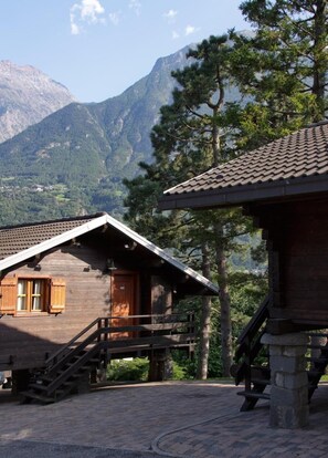 A rustic wooden cabin with a wooden door and windows, a staircase leading to a porch, and a stone bench. - Hôtel Village (Quart)