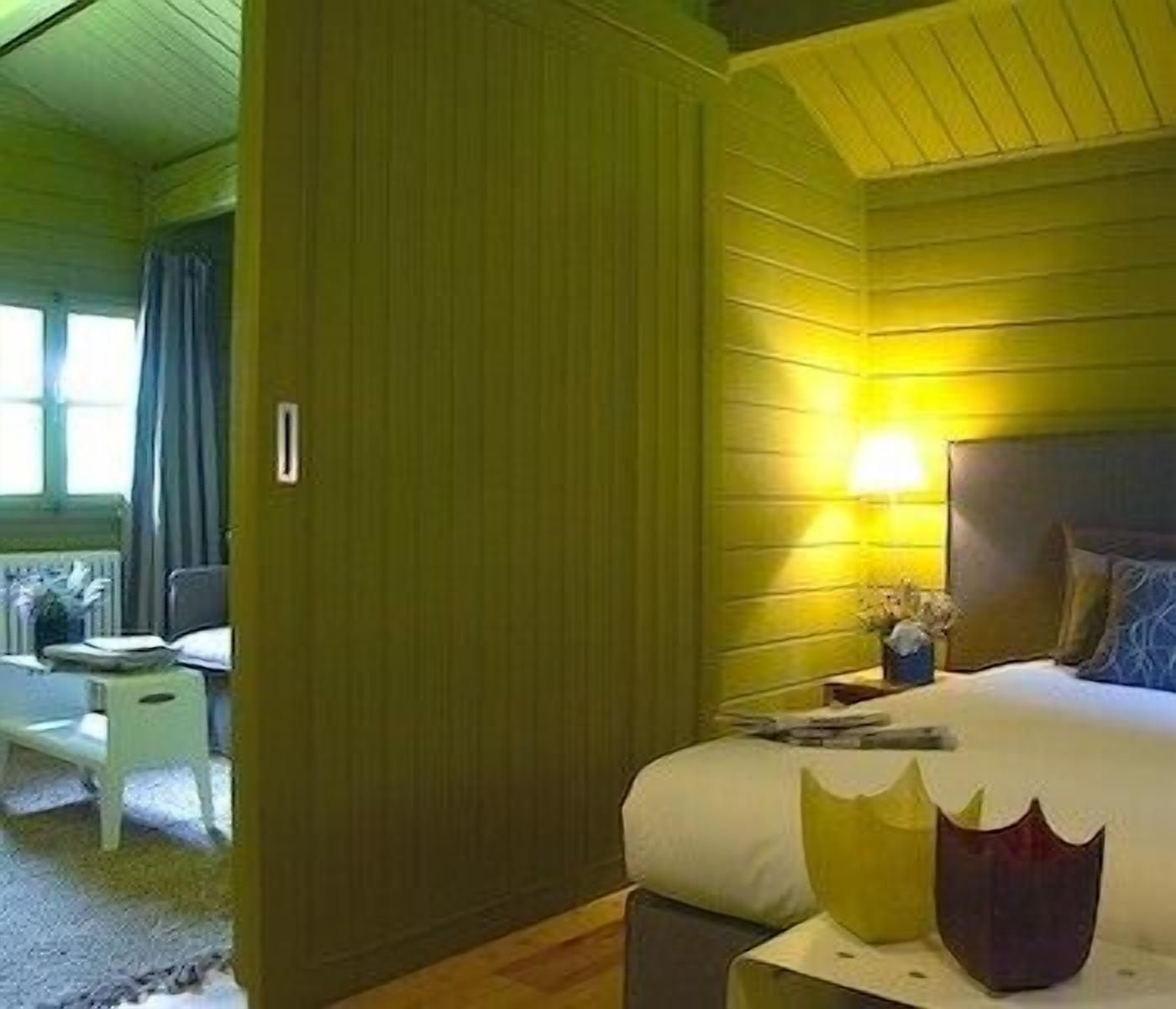 Ein Zimmer mit einem Bett, einem Nachttisch und einer Lampe. Es gibt ein Fenster mit Vorhängen und einen Sessel in der Ecke.