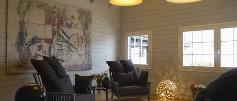 A room with a large abstract painting, two dark armchairs, a round table with a vase of flowers, and a decorative light fixture.