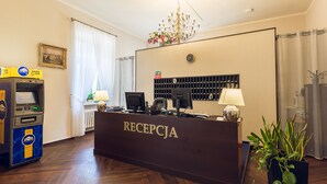 A reception area with a reception desk labeled "RECEPCJA," an ATM, a clock, a plant, and a chandelier. - Hotel Royal (Kraków)
