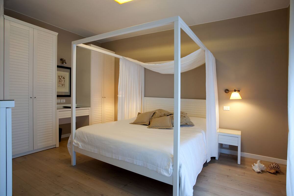 A bedroom with a four-poster bed, a bedside table, a nightstand, a wardrobe, and a wall-mounted light fixture.