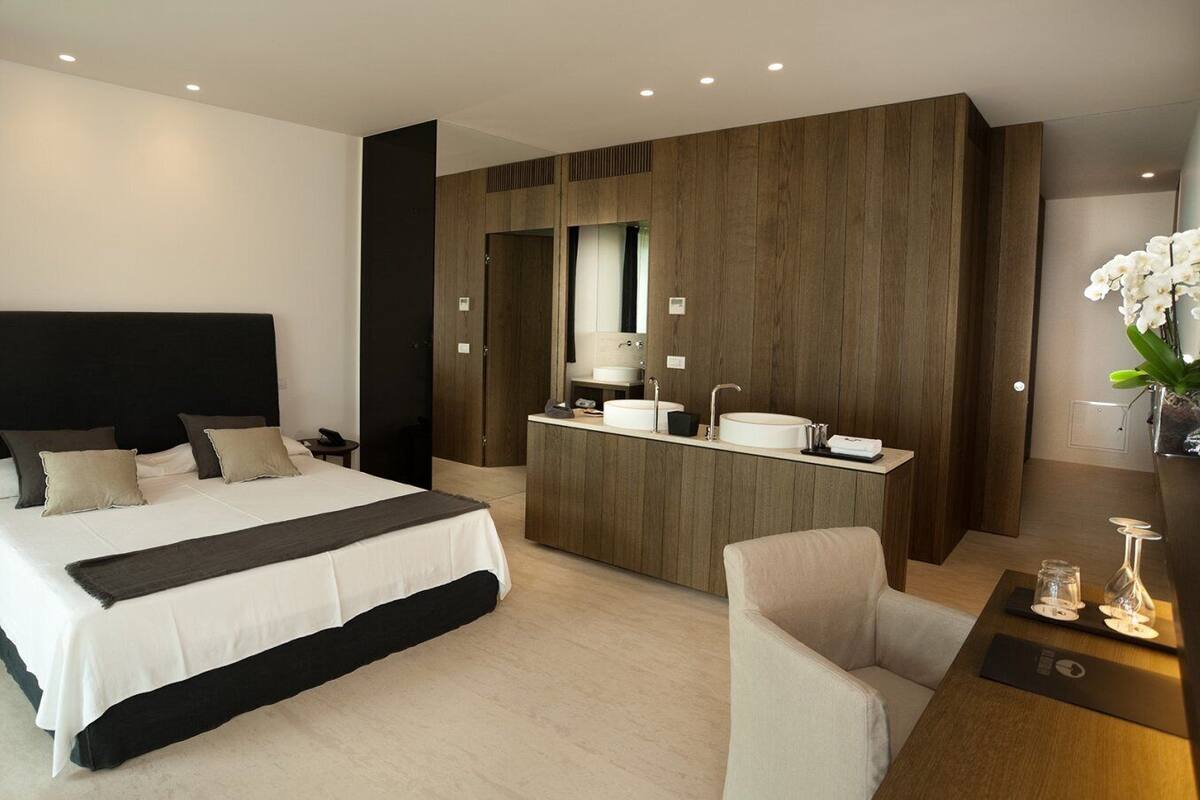 A modern hotel room with a large bed, a small desk, a vanity with a mirror, and a seating area.