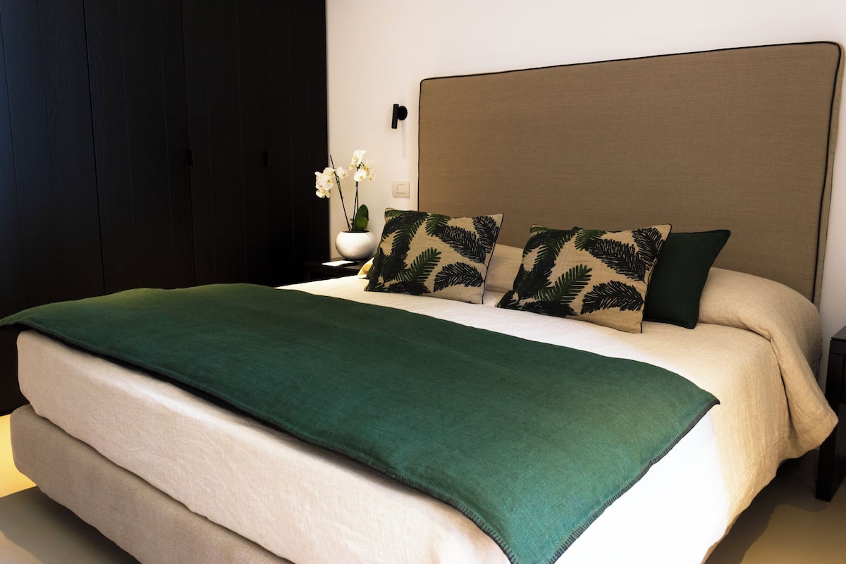 A neatly made bed with a green blanket, two pillows with a leaf pattern, and a vase with white flowers on a bedside table.