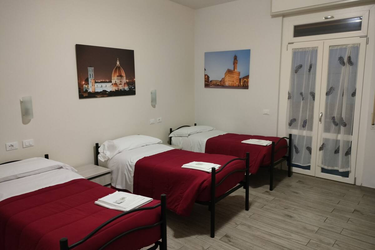 A hotel room with three single beds, each with a red bedspread and white pillows. There are two wall-mounted light fixtures and two framed pictures depicting buildings. The room has a wooden floor and a door with a curtain featuring a feather pattern.