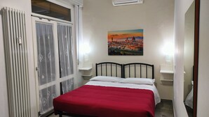 Un dormitorio con cama, un radiador, una ventana con cortinas, aire acondicionado y un cuadro de un paisaje urbano en la pared.