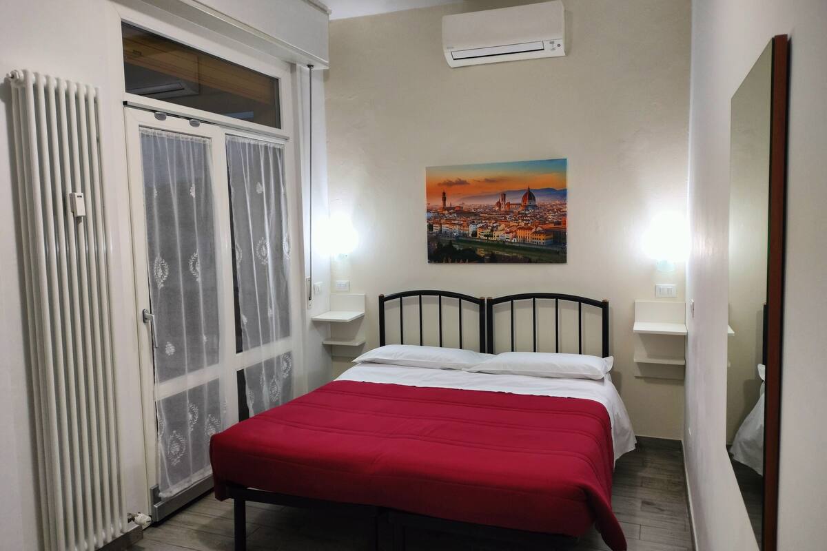 A bedroom with a bed, a radiator, a window with curtains, an air conditioner, and a cityscape painting on the wall.