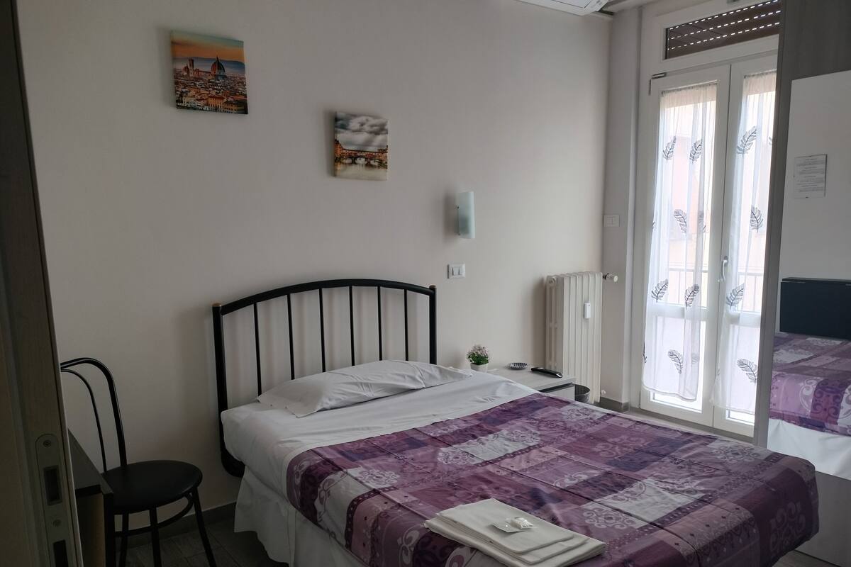 A bedroom with a bed, a chair, a radiator, and two windows with curtains.