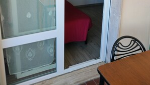 Una habitación con puerta corrediza de vidrio que da a un espacio exterior, una cama con cubrecama rojo, una mesita y una silla.