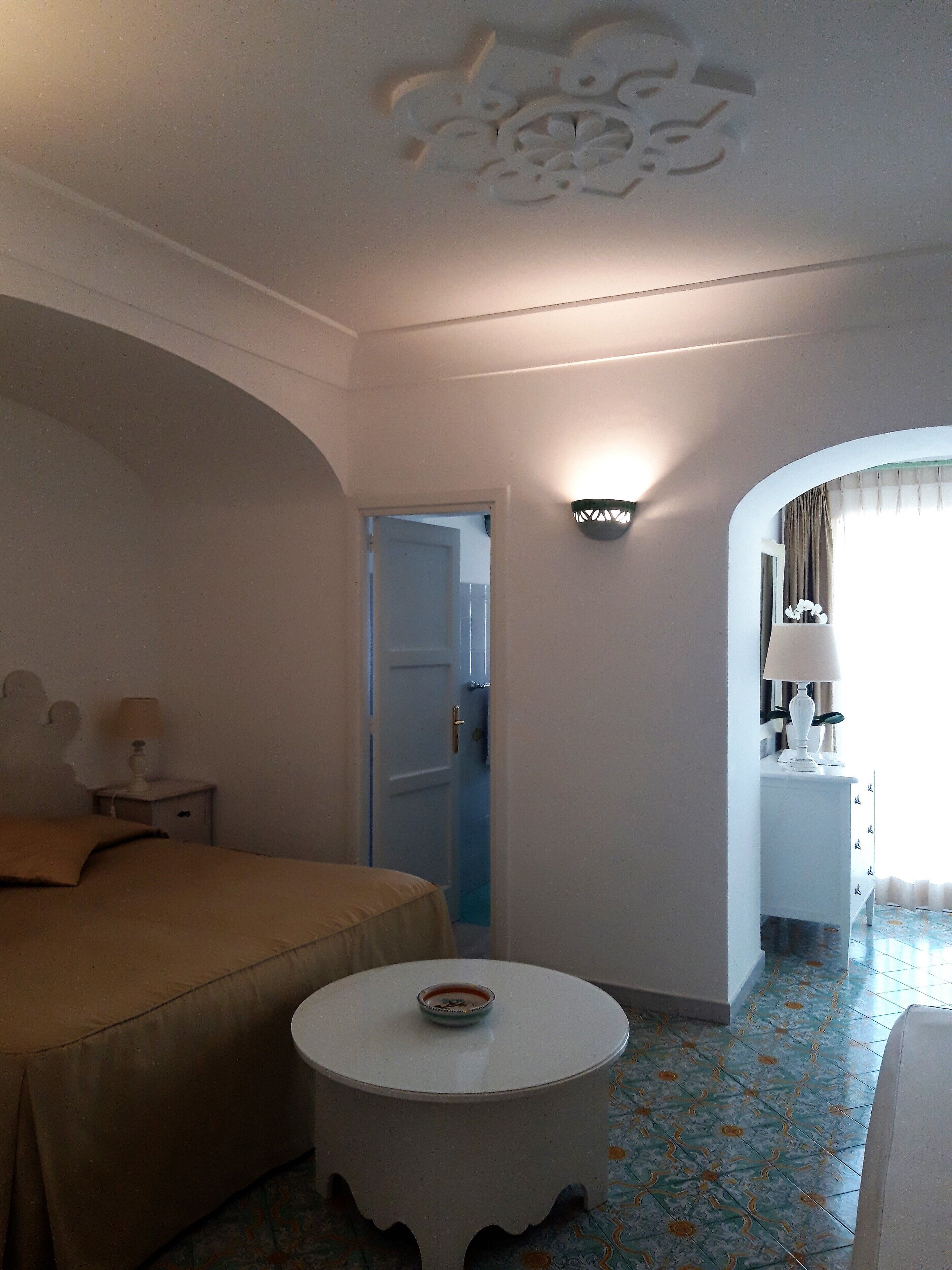 A hotel room with a bed, a round table with a bowl, a nightstand, a lamp, a dresser, and a mirror.