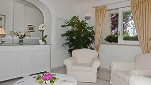 A room with a reception desk, two white armchairs, a large potted plant, and a window with curtains. - Villa Rosa (Positano)