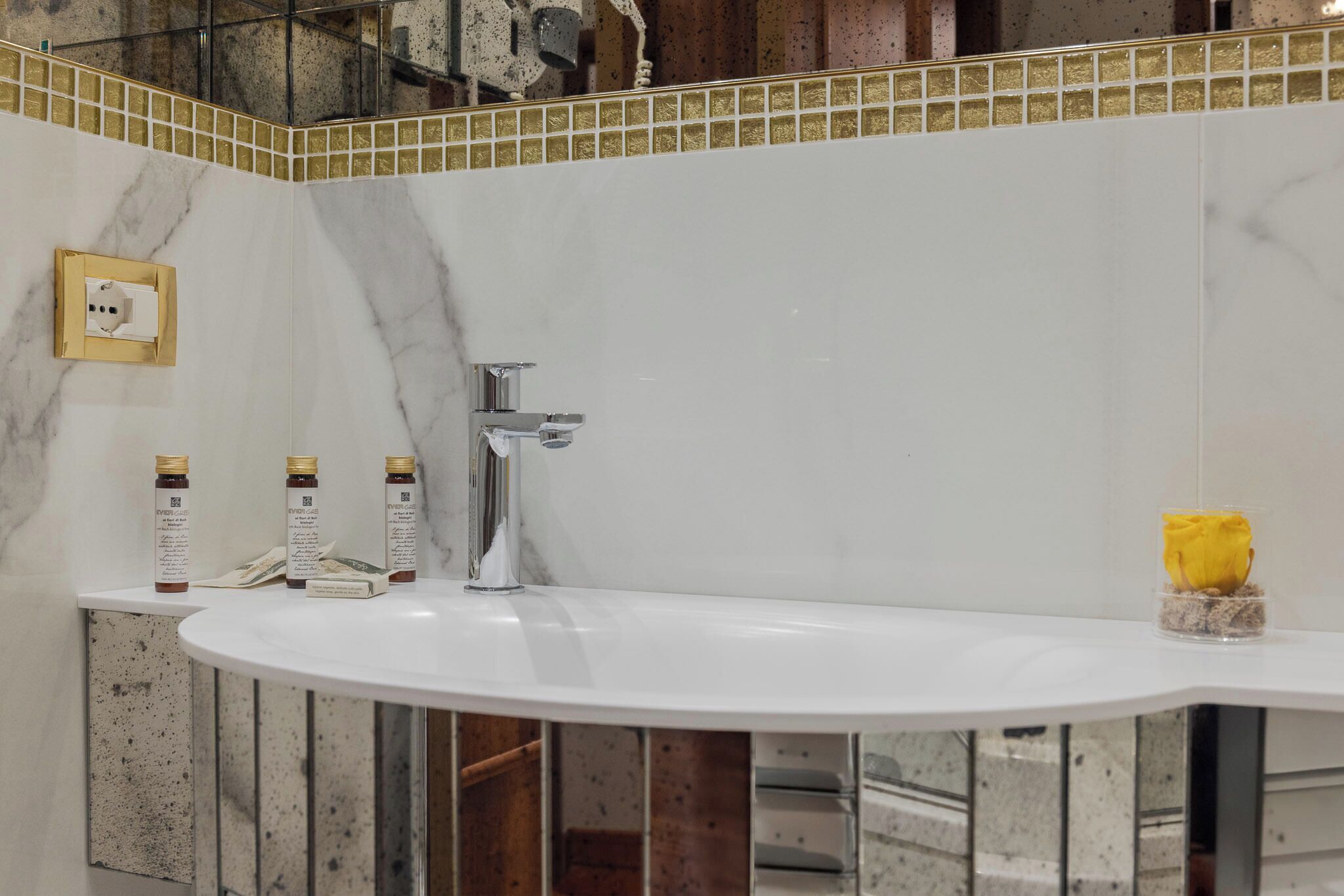 Un baño con un lavamanos blanco, un espejo con marco dorado y una rosa amarilla en un recipiente de vidrio.