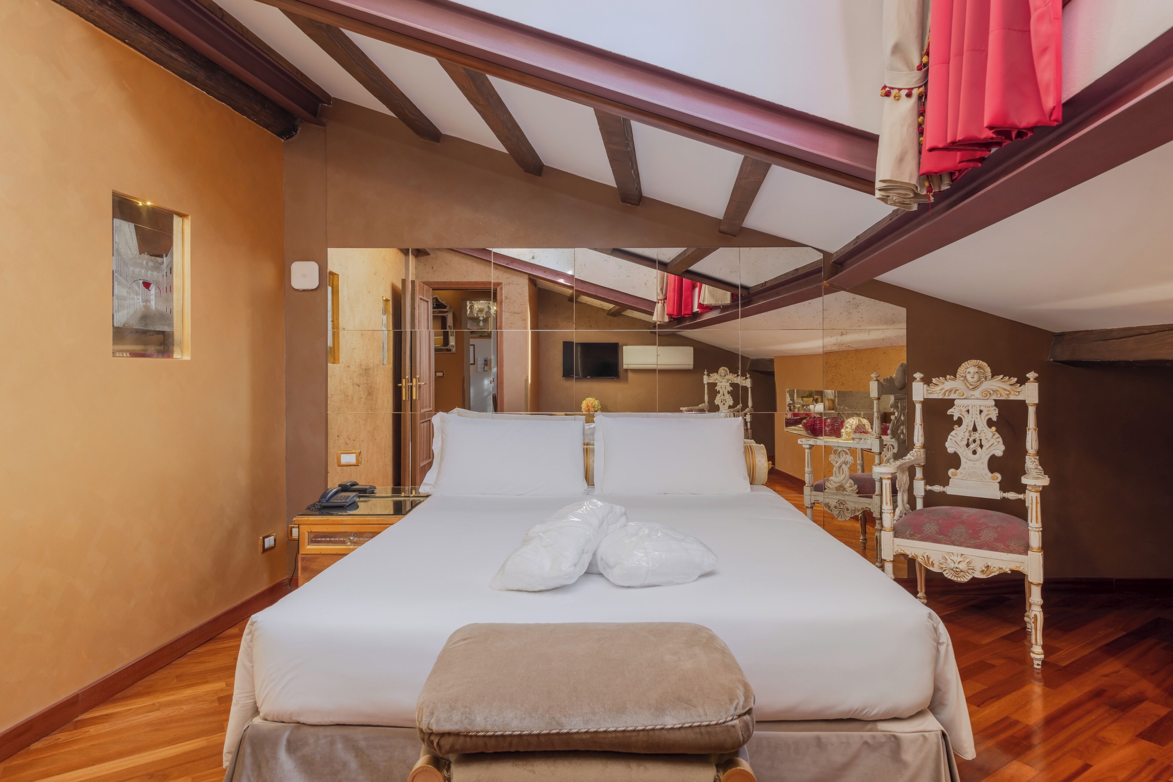 Habitación de hotel con piso de madera, una cama con sábanas blancas, un banco de madera, un espejo y una silla blanca ornamentada.