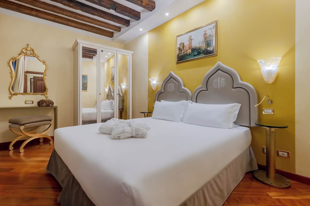 A hotel room with a large bed, a wardrobe, a vanity with a mirror, and a painting on the wall.