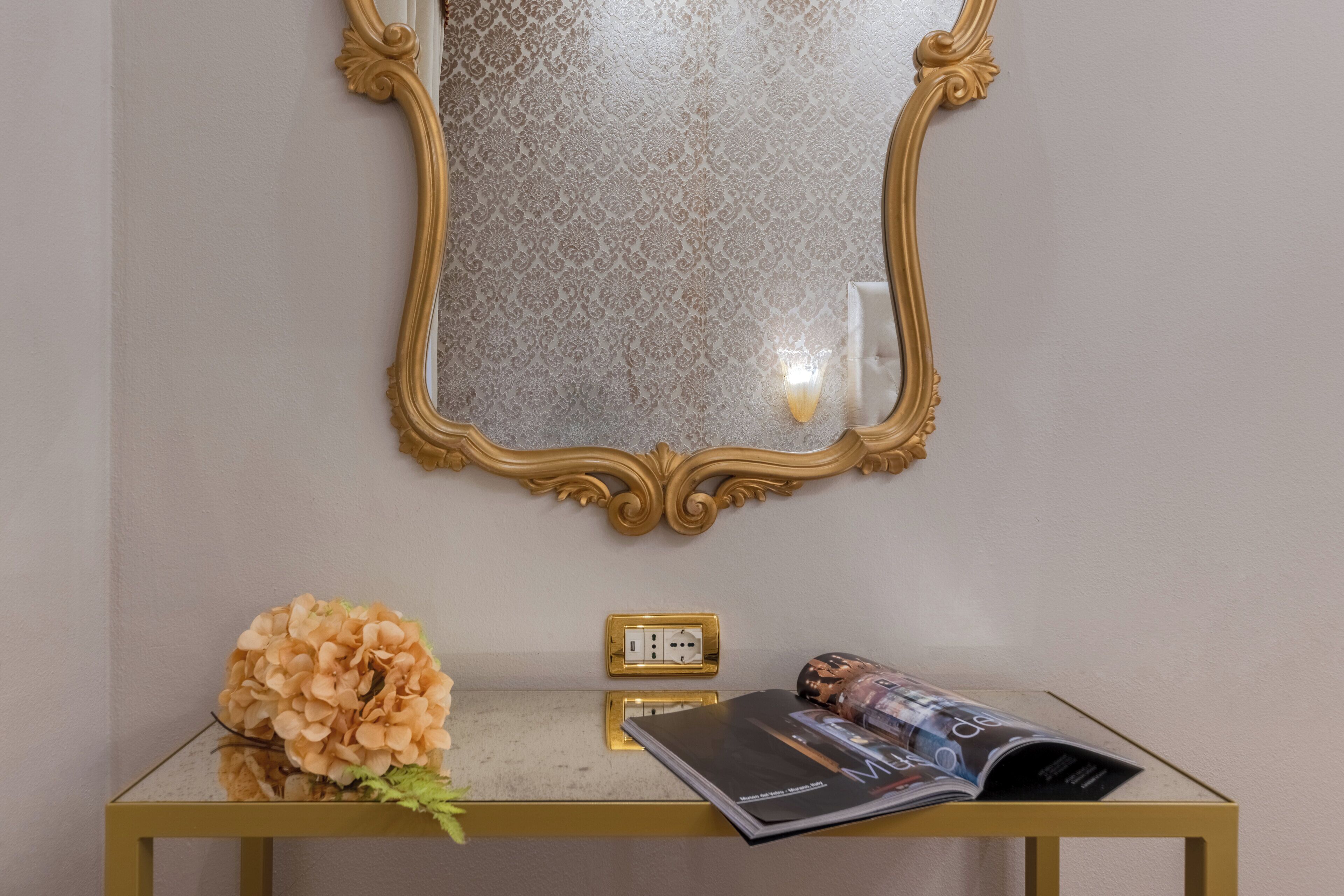 A room with a gold-framed mirror, a glass table with a magazine, and a vase of flowers.