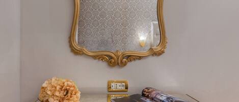 A room with a gold-framed mirror, a glass table with a magazine, and a vase of flowers.